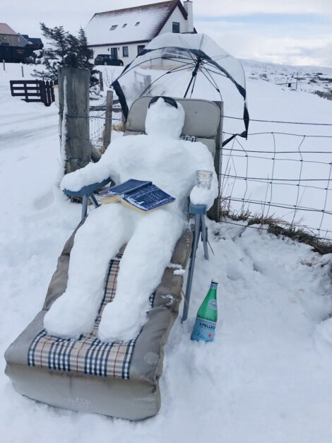 A snowman lies on a reclining chair with an umbrella, holding a drink and a book in a snowy outdoor setting. A bottle of water rests nearby, and a house is visible in the background.