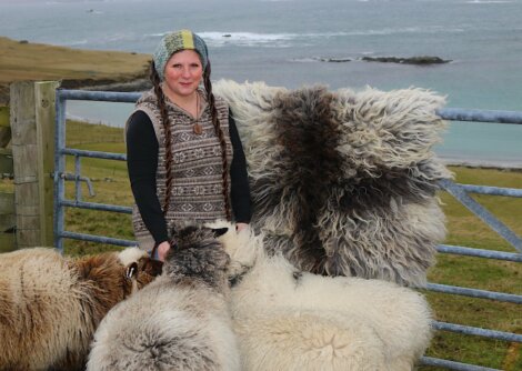 A person stands by a gate near the coast, surrounded by several sheep with varied wool colors. The person is wearing a patterned sweater and a hat, and there is a sheepskin draped over the gate.