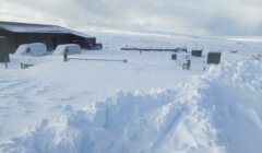 Snow-covered rural area with a barn, vehicles, and fences partially buried in deep snow. Surrounding landscape is also blanketed in snow under an overcast sky.