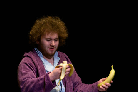 A person in a purple robe is peeling a banana while holding another banana in the other hand against a black background.