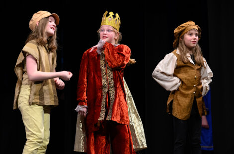 Three children in costume perform on stage. The child in the middle wears a crown and red robe, flanked by two others in peasant-style attire.