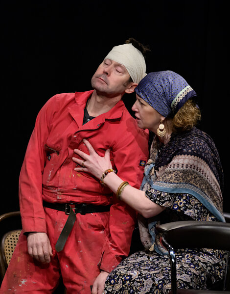 A man in a red jumpsuit with a head bandage appears unconscious, supported by a woman in patterned attire and a headscarf.