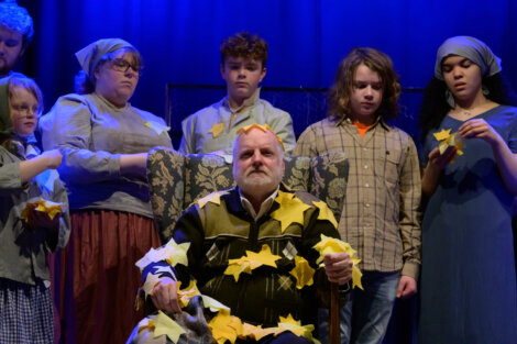 A man sits in a chair covered with yellow paper stars, surrounded by five people who are also holding or wearing yellow stars, set against a dark blue background.