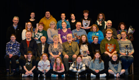 A group photo shows about 30 people of various ages arranged in rows on a stage. Some individuals are holding trophies and awards. All are facing the camera and smiling.