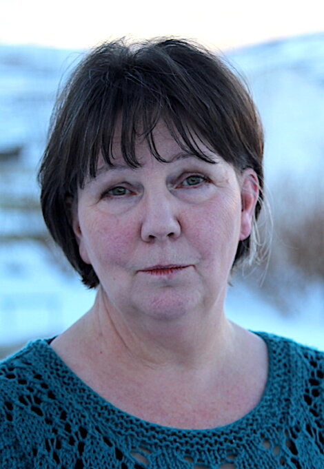 A woman with short dark hair wearing a teal crochet top, standing outdoors with a blurred snowy background. She has a neutral expression on her face.