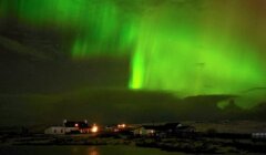 Northern Lights illuminate the night sky with vibrant green hues over a small village with lit houses.