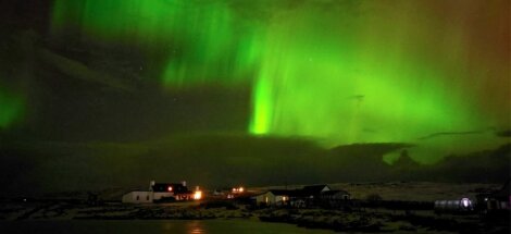 Northern Lights illuminate the night sky with vibrant green hues over a small village with lit houses.