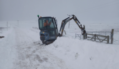 A small excavator is clearing snow on a rural road, creating a path through a thick layer of snow. Snow blankets the surrounding area, and visibility appears reduced due to snowfall.