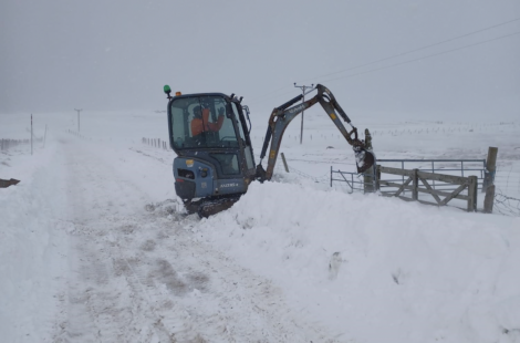 A small excavator is clearing snow on a rural road, creating a path through a thick layer of snow. Snow blankets the surrounding area, and visibility appears reduced due to snowfall.