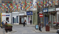 A street scene in a small town with colorful bunting flags overhead. People walk past various shops, and a "No Entry" sign is visible in the foreground.