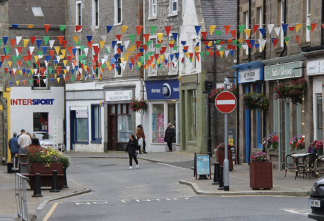 A street scene in a small town with colorful bunting flags overhead. People walk past various shops, and a "No Entry" sign is visible in the foreground.