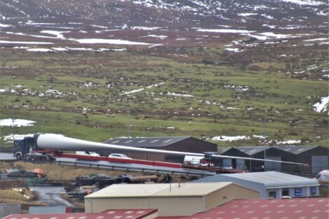 A long truck transports a large wind turbine blade through an industrial area with scattered buildings. Snow patches are visible on a mountainous landscape in the background.