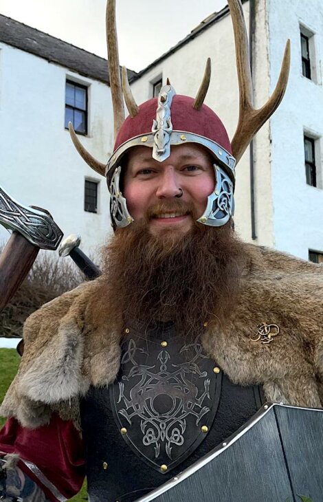 A person dressed in medieval armor, including a horned helmet, chainmail, fur, and holding a sword and shield, stands outside in front of a stone building.