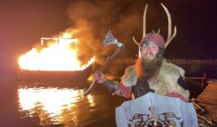 A person dressed in Viking attire stands with an axe and shield, smiling in front of a burning ship on water at night.