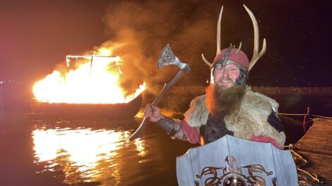 A person dressed in Viking attire stands with an axe and shield, smiling in front of a burning ship on water at night.