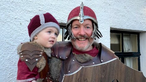 An adult and a child dressed in medieval knight costumes pose in front of a white wall and window. The adult is holding a wooden shield and smiling while the child looks away.