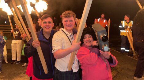 Three people holding torches pose for a photo at an outdoor event. Other participants and a person in a high-visibility vest are in the background.