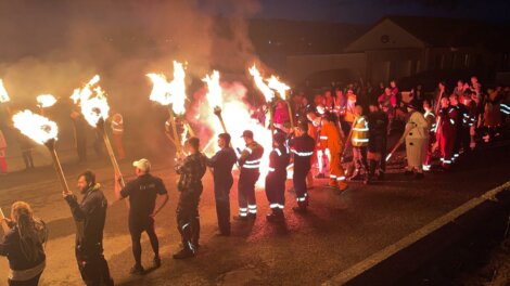 A group of people in reflective clothing holds torches and stands in the street at night, surrounded by a crowd of onlookers.