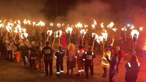 A large group of people, many wearing high-visibility vests, are holding torches with flames at night in an outdoor setting.