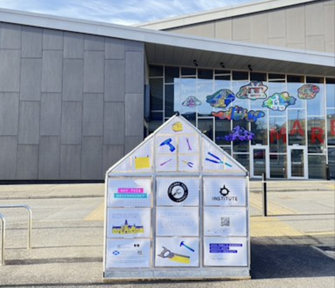 A small, see-through structure with various information panels stands in front of a large building with colorful art above the entrance.