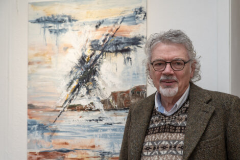 An older man with gray hair and glasses stands in front of an abstract painting. He is wearing a tweed jacket and patterned sweater.