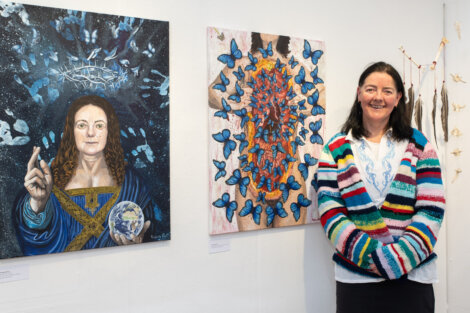 A woman stands beside two paintings, one depicting a person holding a globe with blue butterflies above them, and another showing a figure covered in blue butterflies. She is wearing a striped cardigan.