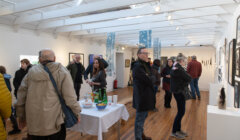 A group of people, some with coats and bags, view artwork and socialize inside a bright gallery space with wooden floors and white walls. A table with snacks and drinks is in the center.