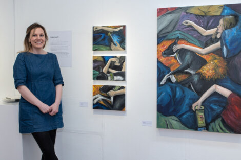 Woman standing next to a set of colorful paintings displayed on a gallery wall, artwork depicts a person and a dog in various positions. A description plaque is mounted on the wall beside her.