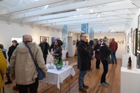 A group of people, some with coats and bags, view artwork and socialize inside a bright gallery space with wooden floors and white walls. A table with snacks and drinks is in the center.