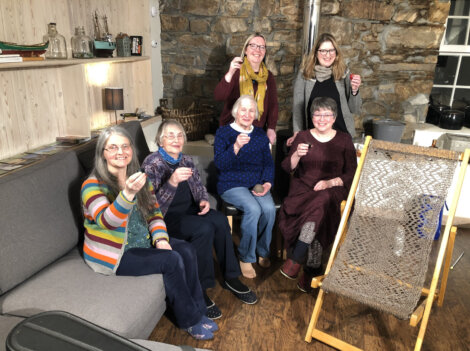 A group of six women, smiling and holding up small glasses, are gathered in a cozy room with wooden and stone walls. A loom is visible in the foreground.