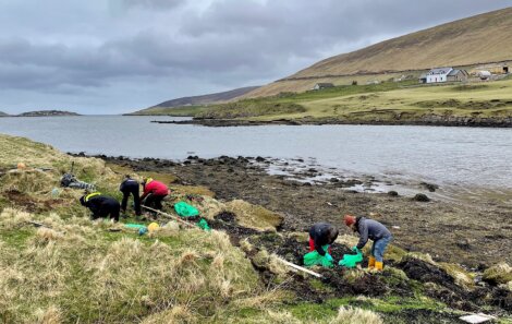 Four people in outdoor clothing clean up debris along a rocky shoreline with grassy hills and houses in the background on a cloudy day.