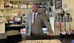 A smiling man in a checked suit stands behind a well-stocked bar with a cash register, bottles of various liquors, and beer taps. The bar features a sign that reads "The Adventure Begins Here.