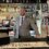 A smiling man in a checked suit stands behind a well-stocked bar with a cash register, bottles of various liquors, and beer taps. The bar features a sign that reads "The Adventure Begins Here.