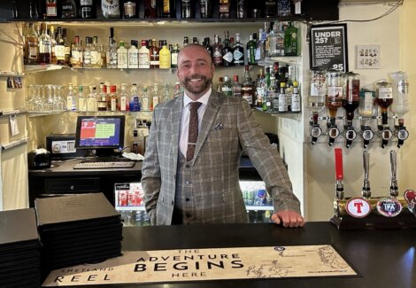 A smiling man in a checked suit stands behind a well-stocked bar with a cash register, bottles of various liquors, and beer taps. The bar features a sign that reads "The Adventure Begins Here.