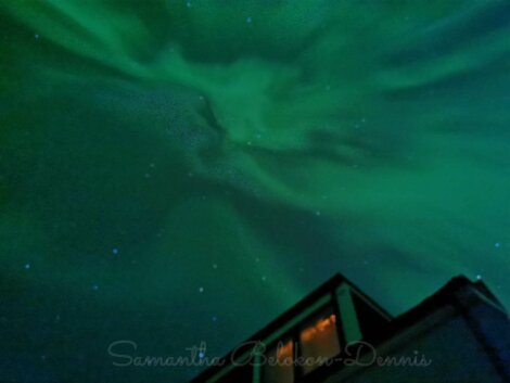 Night sky illuminated by green and blue auroras, with a part of a building and a window visible in the lower right corner.