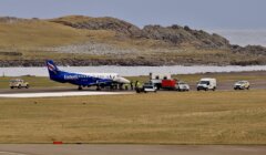 A small passenger plane with "Eastern Airways" livery is stationed on a runway, surrounded by several utility vehicles and personnel, with a grassy landscape and rocky hills in the background.