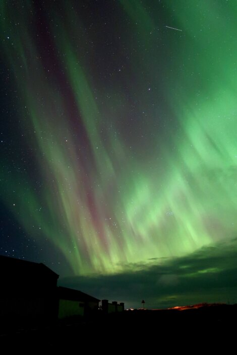 The image shows the Northern Lights, with green and subtle red hues, illuminating a star-filled night sky. A structure is silhouetted in the bottom left corner.