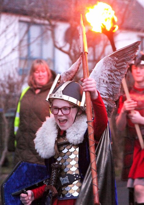 A person dressed in medieval armor with wings, a helmet, and a fur mantle holds up a staff with a flame in the background during an outdoor event.