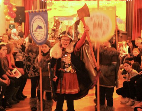 A child in a Viking costume, holding an axe and shield, stands excitedly at the center of a crowded indoor event while other children and spectators look on. Banners are displayed in the background.