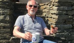 A man with sunglasses and a grey polo shirt sits on a stone structure, holding a white mug and a piece of food.