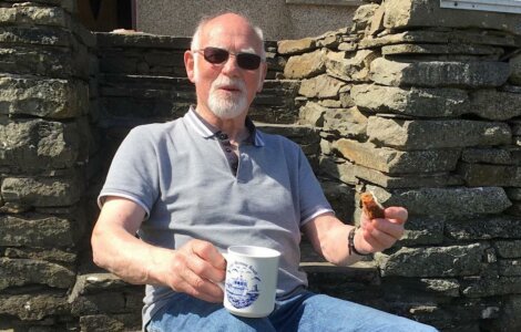 A man with sunglasses and a grey polo shirt sits on a stone structure, holding a white mug and a piece of food.