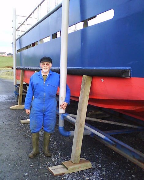 A person in a blue coverall and green boots stands next to a large blue and red boat that is supported by wooden braces.