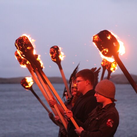 People standing in a line holding lit torches over a body of water with a cloudy sky in the background.