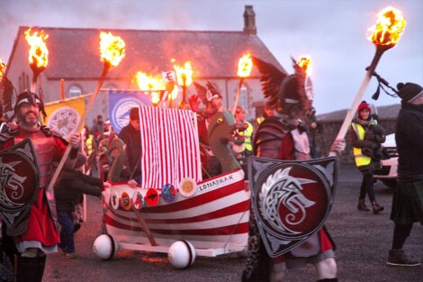 A group of people in Viking costumes parading with a small Viking ship float, holding lit torches and shields, during an outdoor event in front of a stone building.