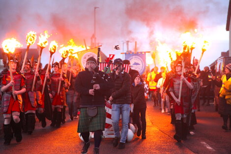 People parade with fire torches and banners in a dusk setting. A person dressed in a green tartan kilt plays the bagpipes while others, including participants in Roman attire, follow.