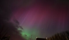 A night sky displays colorful aurora borealis with shades of green and pink above a silhouetted house and bare trees.