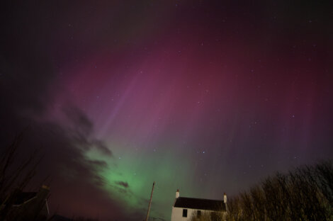 A night sky displays colorful aurora borealis with shades of green and pink above a silhouetted house and bare trees.