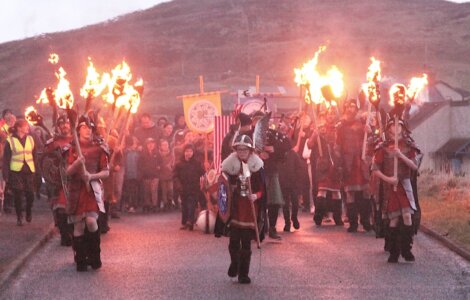 A group dressed in traditional Viking attire parades, carrying flaming torches and shields, followed by a crowd. The scene takes place on a road with a hill and houses in the background.