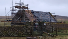 A stone building is under renovation, covered in scaffolding and a tarp. The scene includes a stone wall, a gate, and grassy surroundings with a cloudy sky overhead.