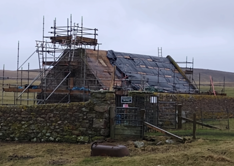 A stone building is under renovation, covered in scaffolding and a tarp. The scene includes a stone wall, a gate, and grassy surroundings with a cloudy sky overhead.